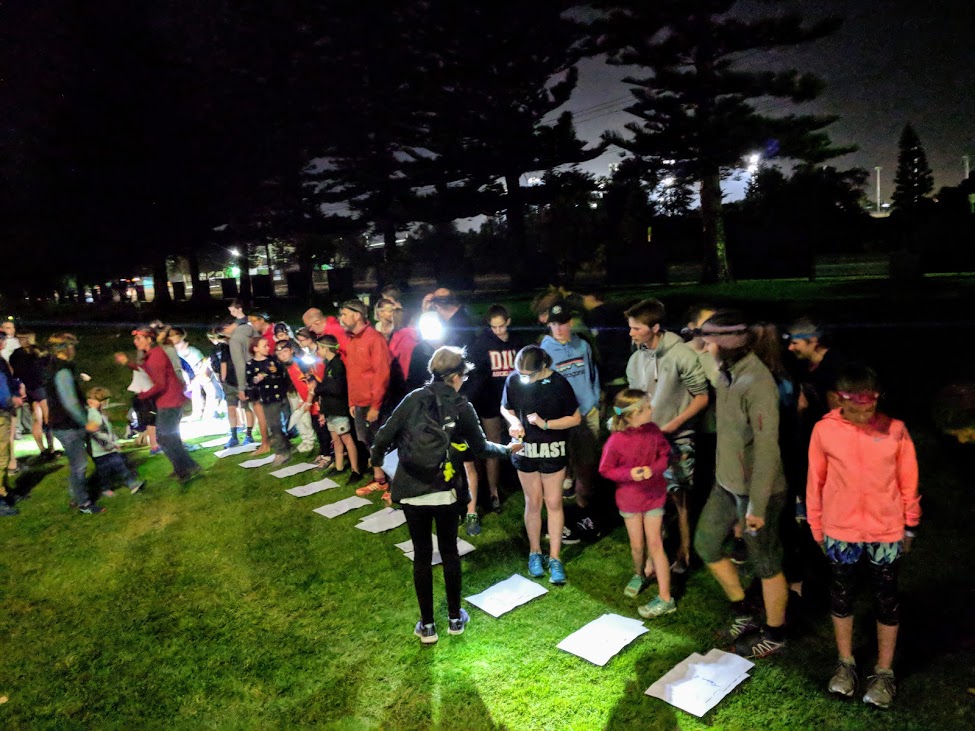 Group of people, mostly teens, gathered on a lit grassy area at night with papers laid out on the ground as part of an activity surrendering clues or tasks.