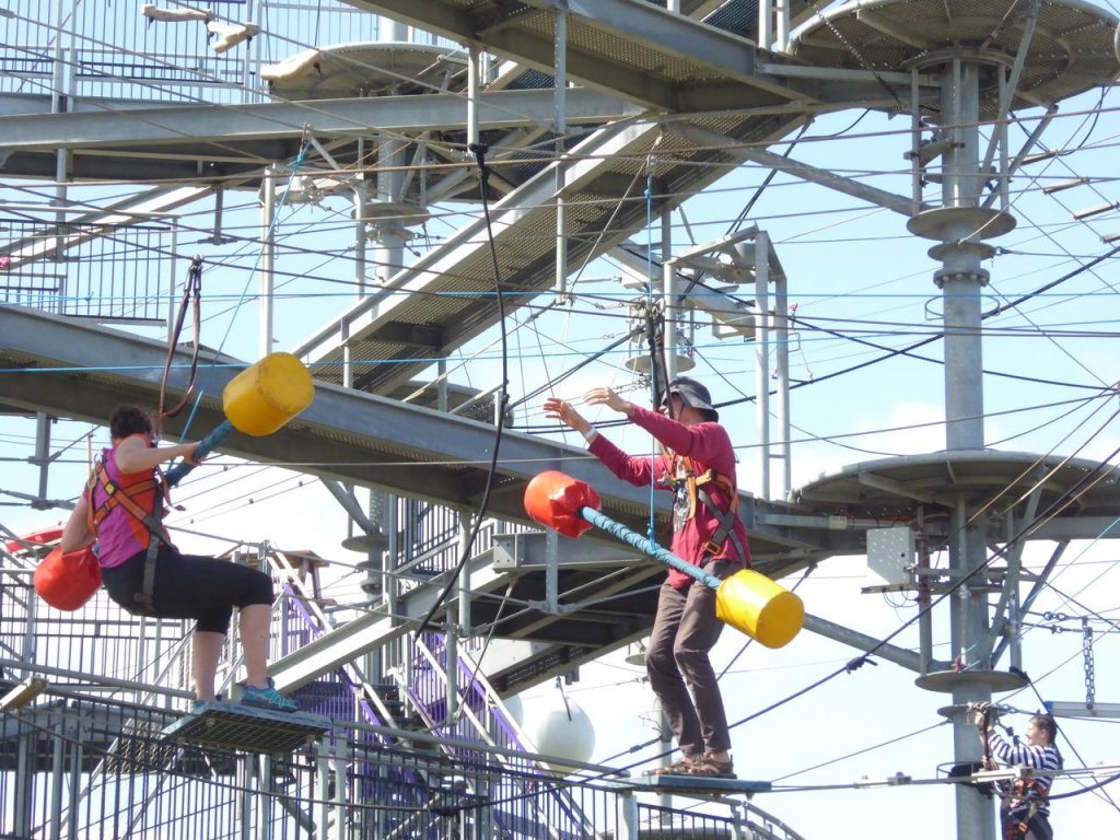 Two people in safety harnesses navigate a metal high-ropes obstacle course outdoors, reaching toward each other amid cables and platforms.