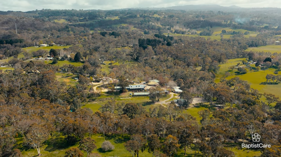 Aerial view of a wooded rural landscape with scattered houses, fields, and winding roads; BaptistCare logo in the bottom-right corner.