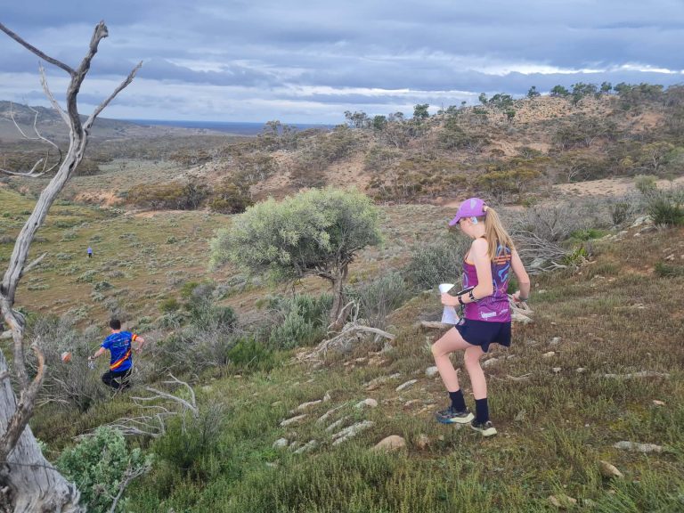 Runners traverse a rocky desert trail with scrubby bushes and distant hills under a cloudy sky; a woman in purple running gear and a pink cap is in the foreground.